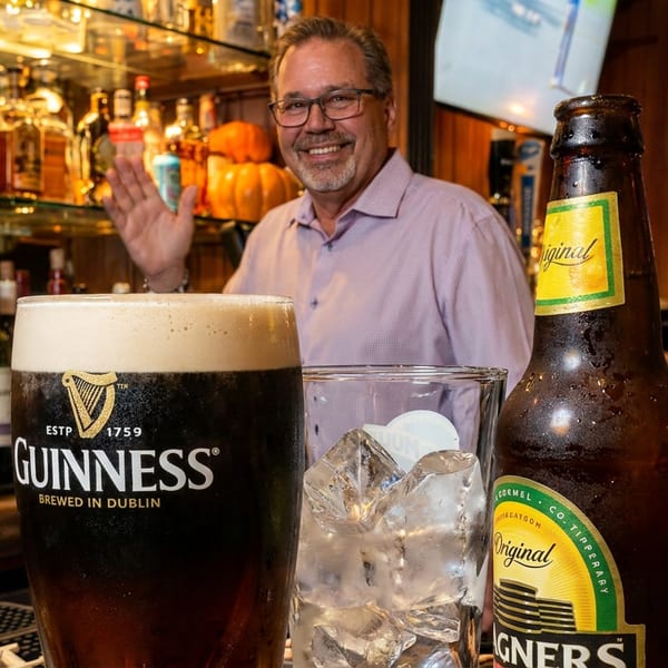 A smiling man waves from behind a bar with a Guinness, a glass of ice, and a beer bottle.