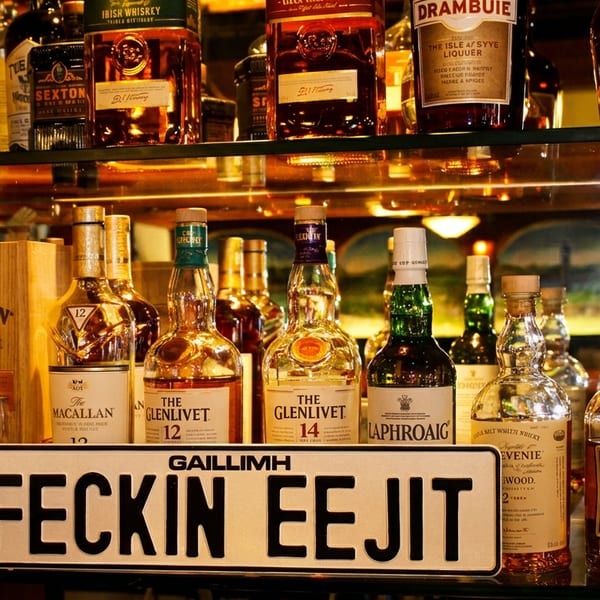 A bar shelf filled with various whiskey and liquor bottles, with a "FECKIN EEJIT" sign in front.