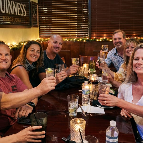 Eight smiling people toast with drinks around a candlelit table in a warmly lit restaurant.