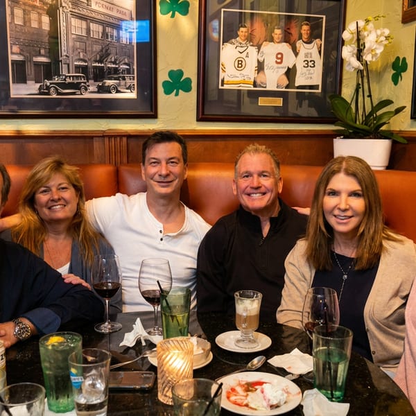 Six smiling people, three men and three women, seated at a restaurant booth with drinks and dessert.