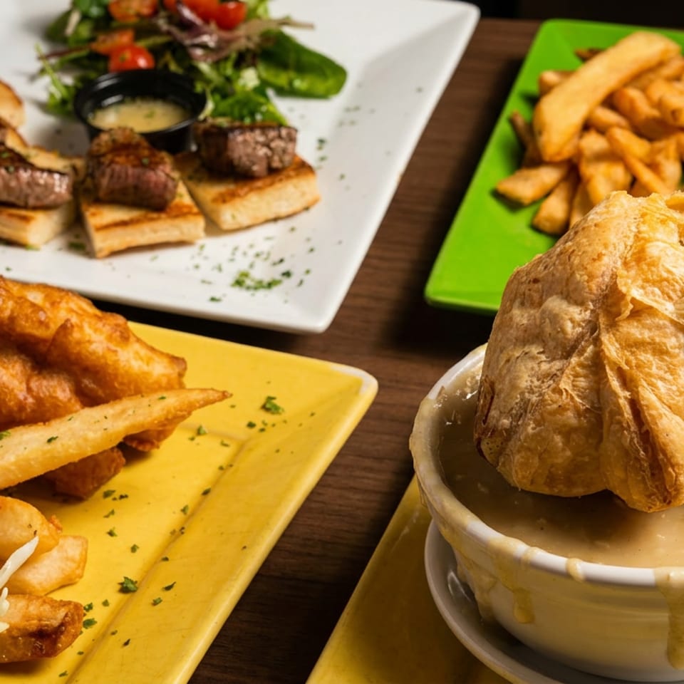A table spread with fish and chips, steak bites, a burger with fries, and a soup with a pastry topping.