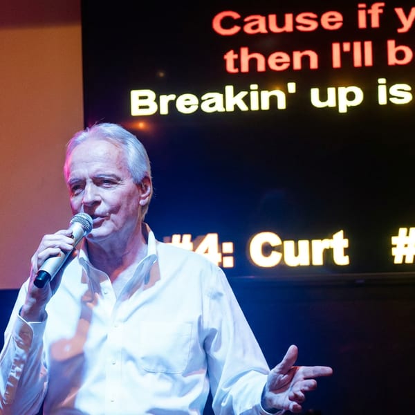 Man in a white shirt sings into a microphone onstage beneath colorful karaoke lyrics and rankings.